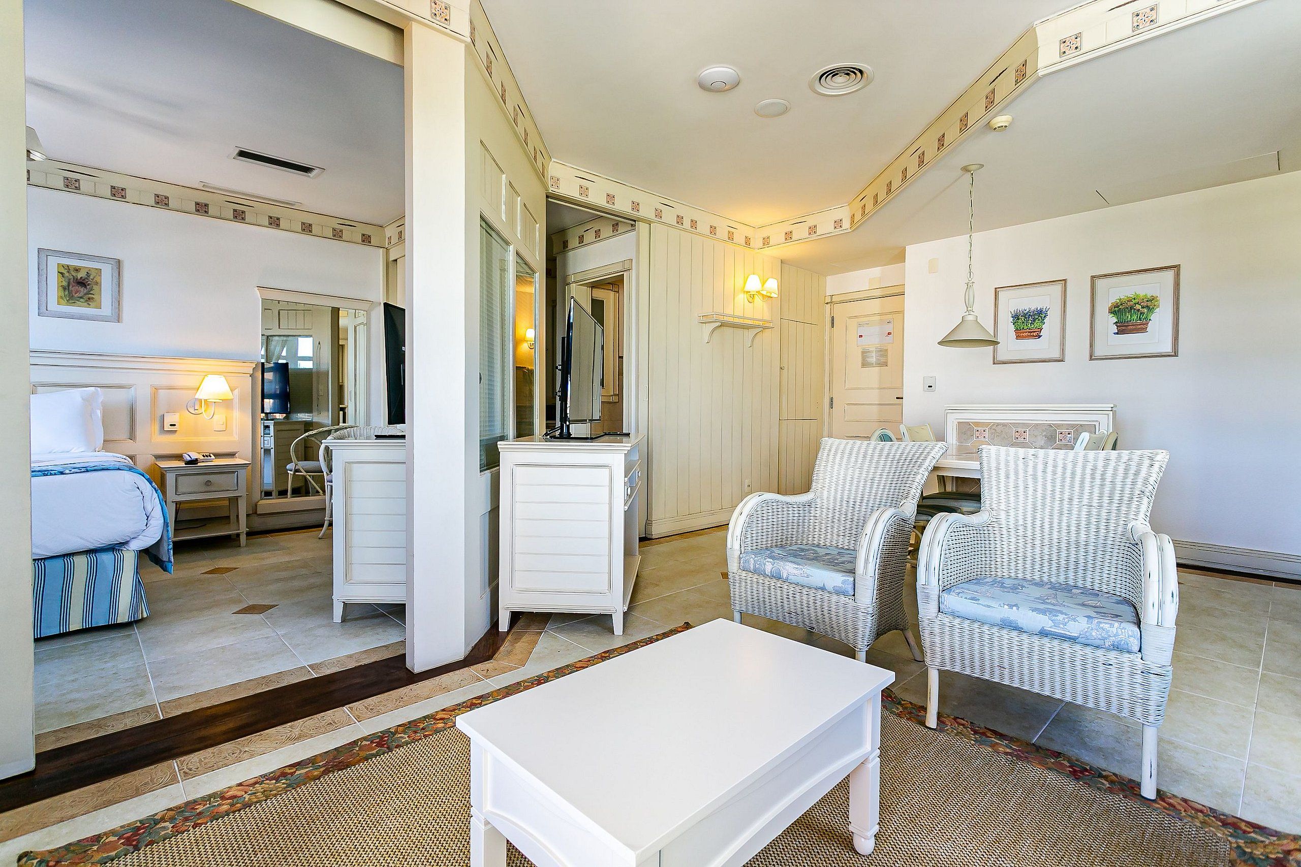 A hotel room with a bed, a white dresser, two wicker chairs, a white coffee table, a mirror, and a small dining area.