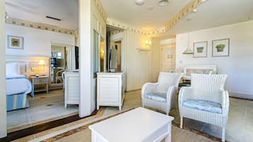 A hotel room with a bed, a white dresser, two wicker chairs, a white coffee table, a mirror, and a small dining area.