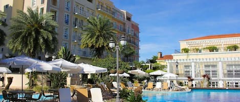 A hotel pool area with multiple lounge chairs and umbrellas, surrounded by palm trees and a large building in the background.