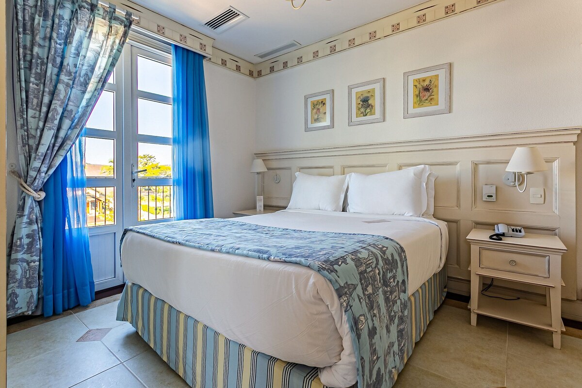 A neatly arranged bedroom with a large bed, a bedside table with a lamp, and a window with curtains.
