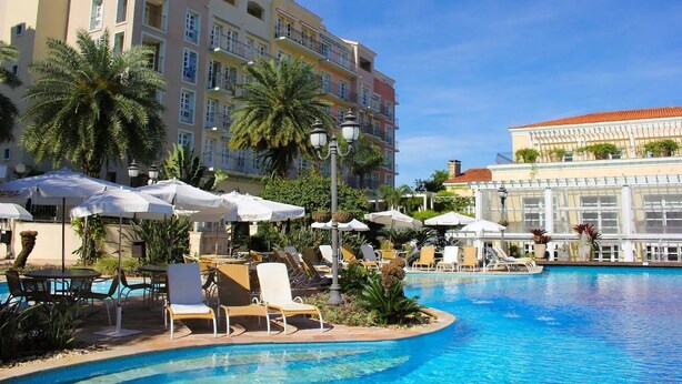 A hotel pool area with multiple lounge chairs and umbrellas, surrounded by palm trees and a large building in the background. - Studios frente ao mar no IL Campanario Resort ILCTOP (Florianópolis)