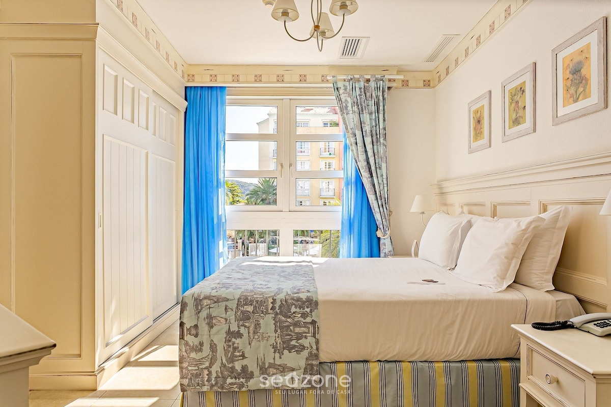 Una habitación de hotel con cama, mesita de noche, teléfono y ventana con cortinas.