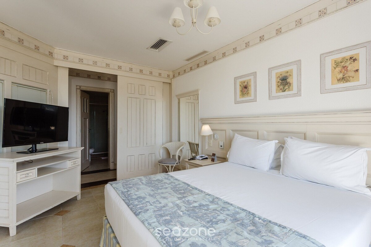Una habitación de hotel con una cama, un televisor sobre una cómoda blanca, una silla y una mesita de noche con una lámpara.