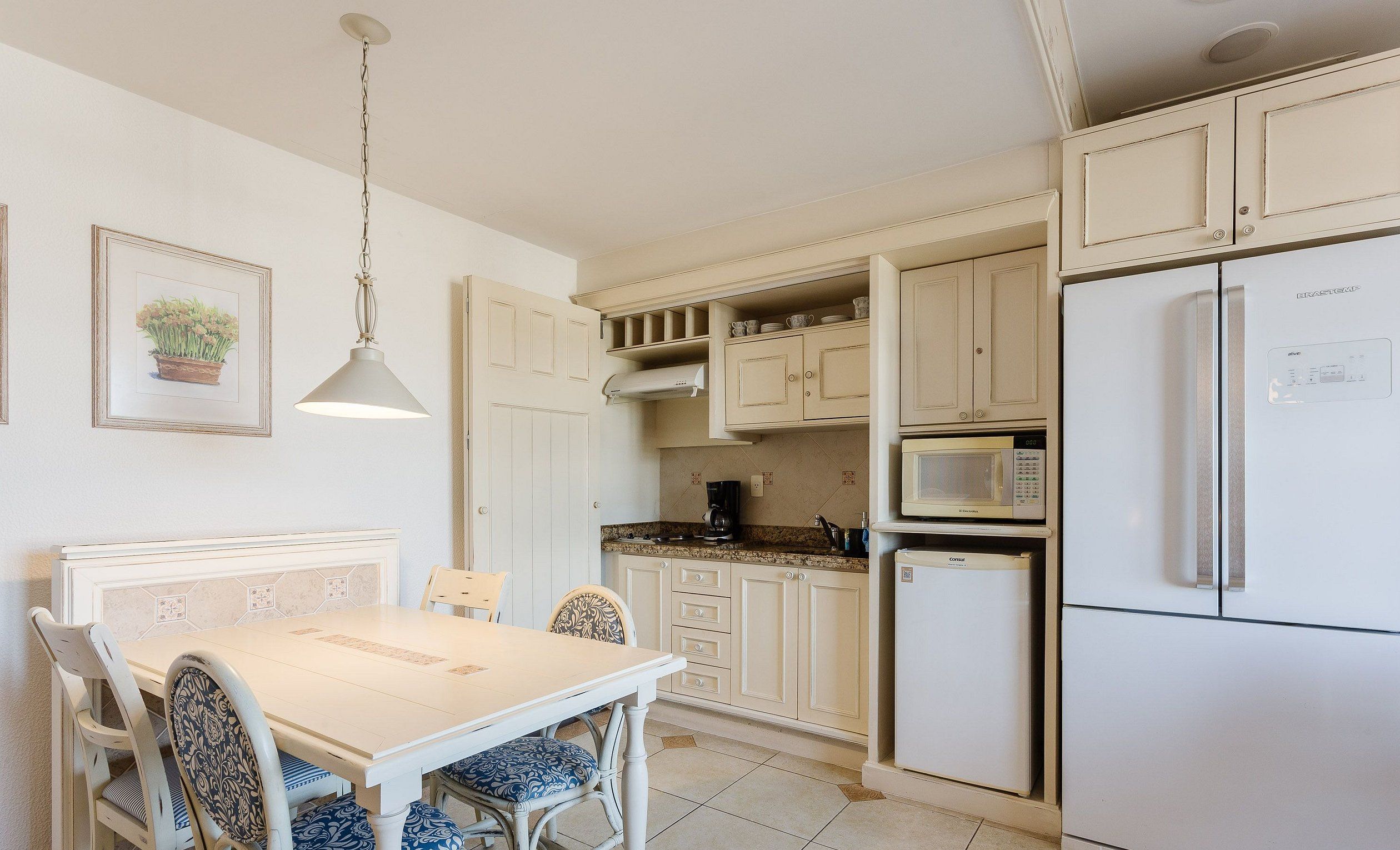 Una cocina con gabinetes blancos, una mesa de comedor con sillas, una lámpara colgante y un refrigerador.