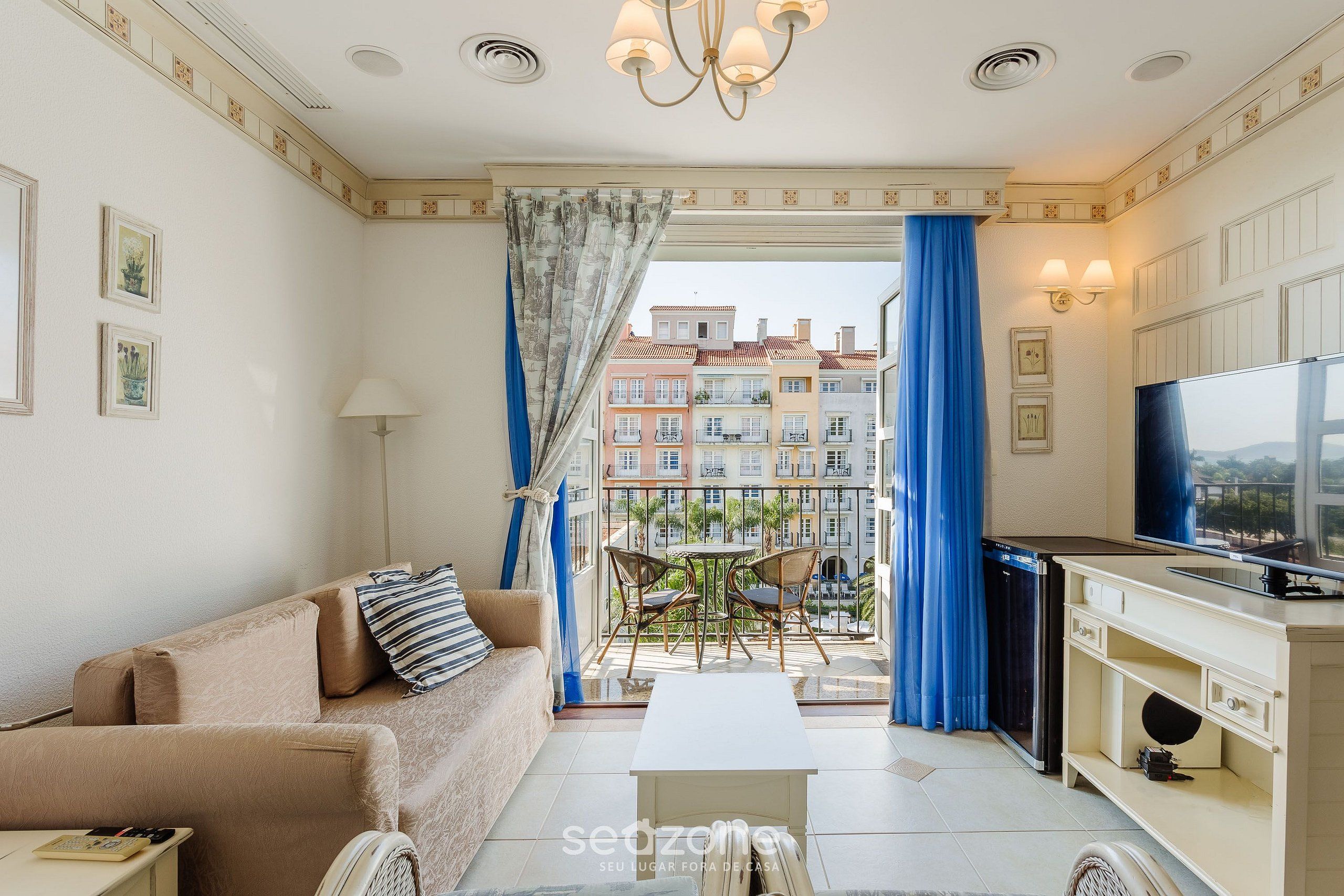 Una sala moderna con sofá, mesa de centro y comedor. Se ve un balcón con cortinas azules y vistas a edificios.