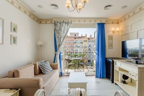 A modern living room with a sofa, coffee table, and dining area. A balcony with blue curtains and a view of buildings is visible. - Studios frente ao mar no IL Campanario Resort ILCTOP (Florianópolis)