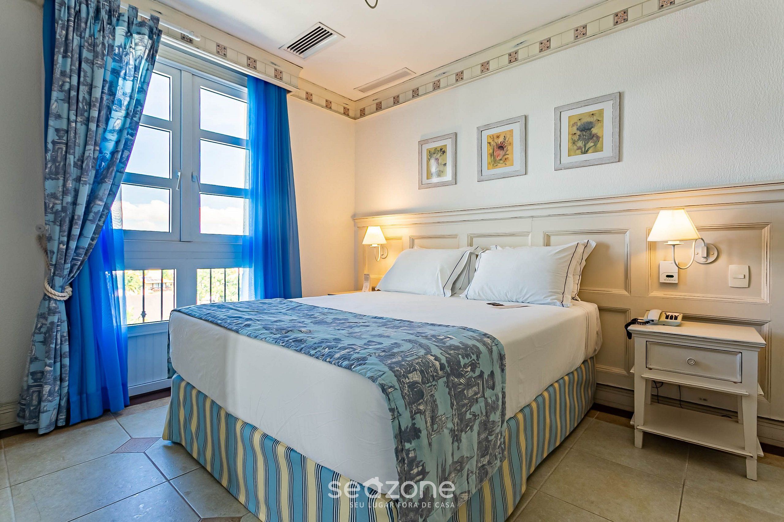 Una habitación de hotel con una cama grande, una mesita de noche con lámpara y una ventana con cortinas azules.