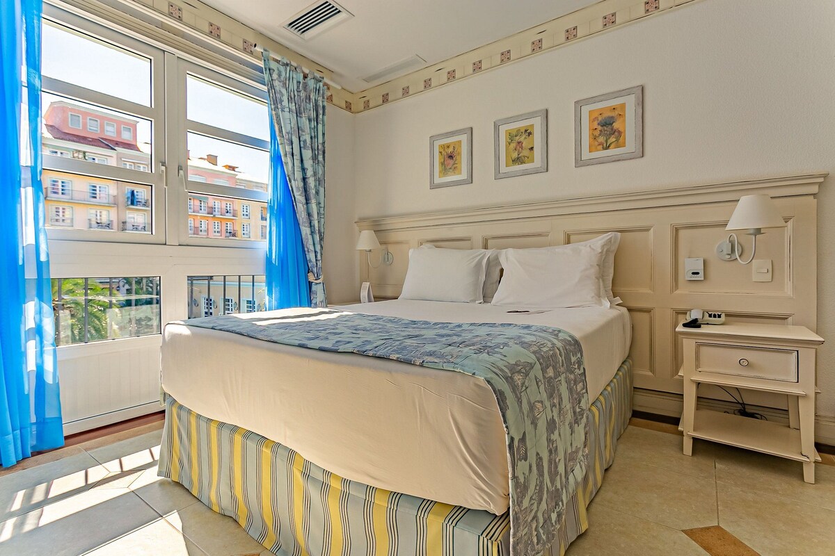 Habitación de hotel con cama, mesita de noche, ventana con cortinas azules y vista a edificios.