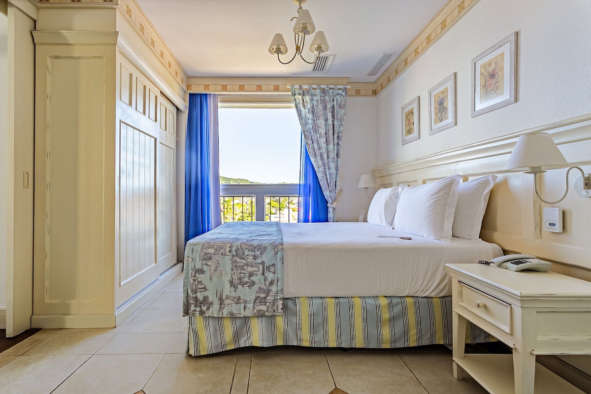 Una habitación de hotel con una cama grande, una mesita de noche con lámpara, una ventana con cortinas azules y cuadros en la pared.
