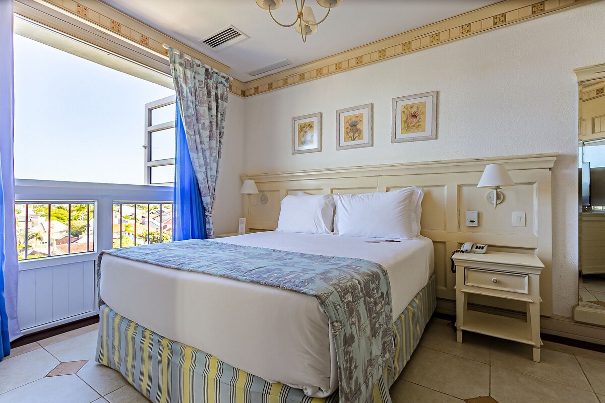 Una habitación de hotel con cama, mesita de noche, lámpara y ventana con cortinas.