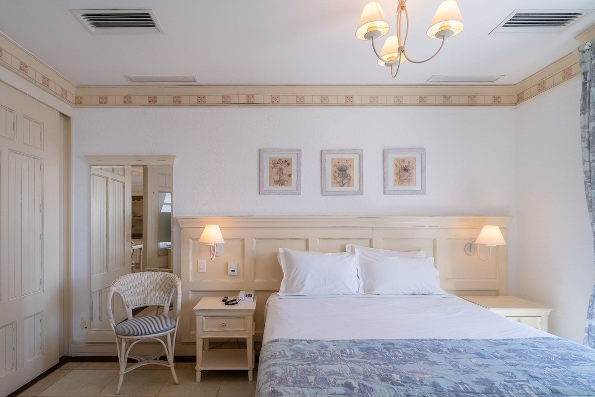 A hotel room with a bed, a nightstand, a chair, a wardrobe, and a chandelier.