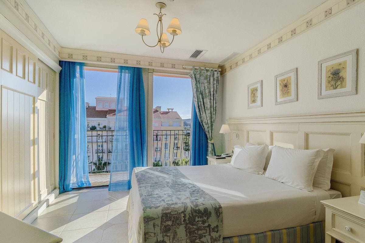 Habitación de hotel con una cama grande, una mesita de noche, un candelabro y vistas a edificios a través del ventanal.