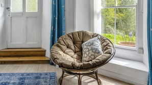 Een kamer met een groot raam, een deur met geëtste ruitpanelen, een bruine fauteuil met een geknoopt kussen, en een blauw vloerkleed.