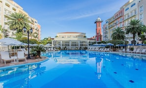 Une grande piscine intérieure entourée de chaises longues et de parasols, avec un bâtiment doté d’une tour en arrière-plan.