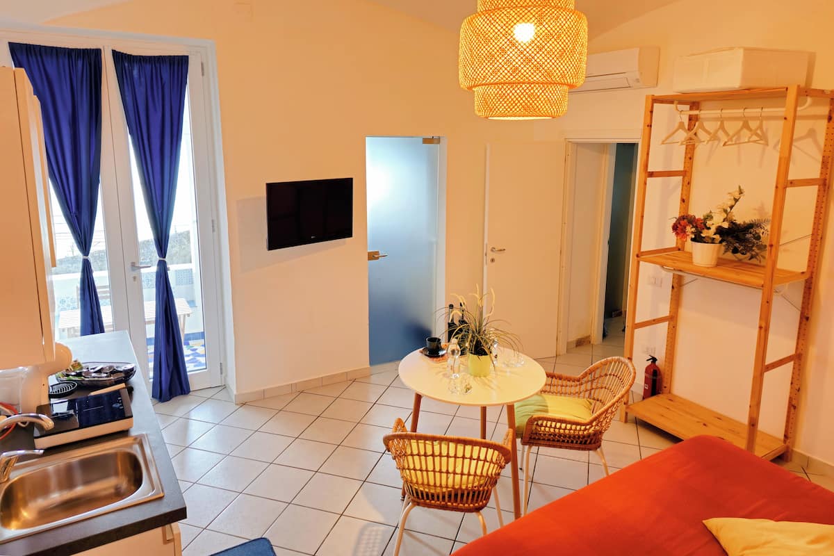 Ein kompakter Wohnraum mit Essbereich, einem an der Wand befestigten Fernseher, einem Holzregal mit Pflanze und einem roten Sofa.