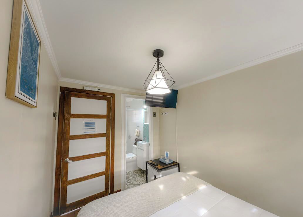 A hotel room with a bed, a bedside table, a hanging light fixture, a door, and a bathroom visible through an open door.