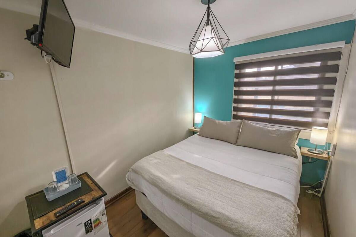 Habitación de hotel con cama, televisor montado en la pared, una mesita de noche con lámpara y ventana con persianas.