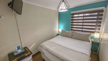 Habitación de hotel con cama, televisor montado en la pared, una mesita de noche con lámpara y ventana con persianas.