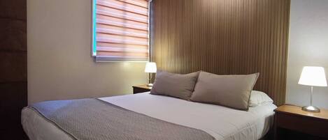 A hotel room with a large bed, wooden paneling on the walls, a window with blinds, and two bedside lamps.