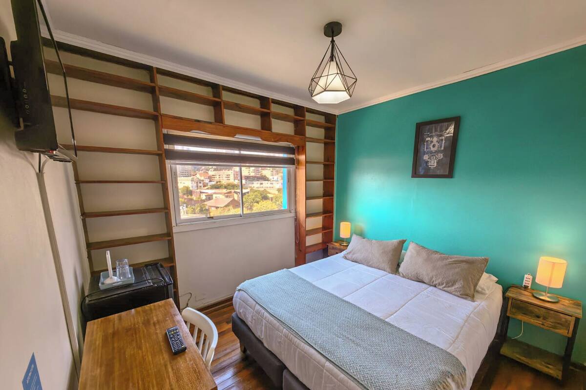 Un dormitorio con cama, mesitas de noche, un escritorio, una silla, una ventana con vistas y una lámpara colgante.