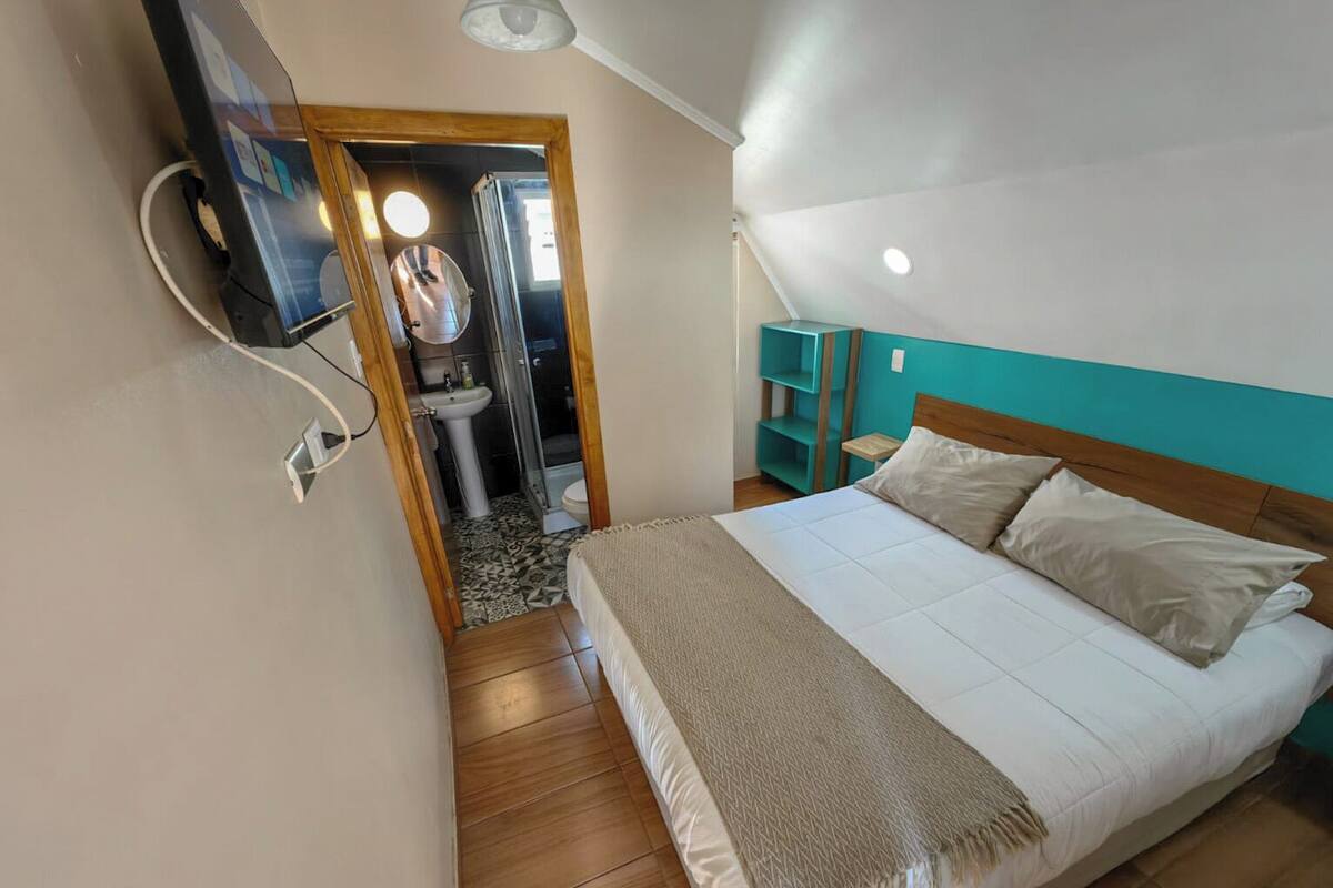 Un dormitorio con cama, mesita de noche, espejo y baño visible a través de una puerta abierta.