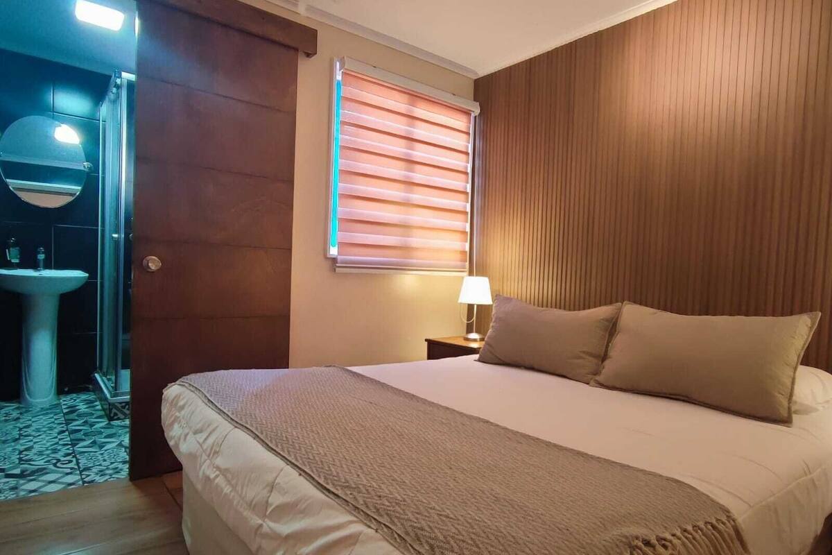 Un dormitorio con una cama grande, una mesita de noche con lámpara, una ventana con persianas y un baño visible a través de una puerta abierta.