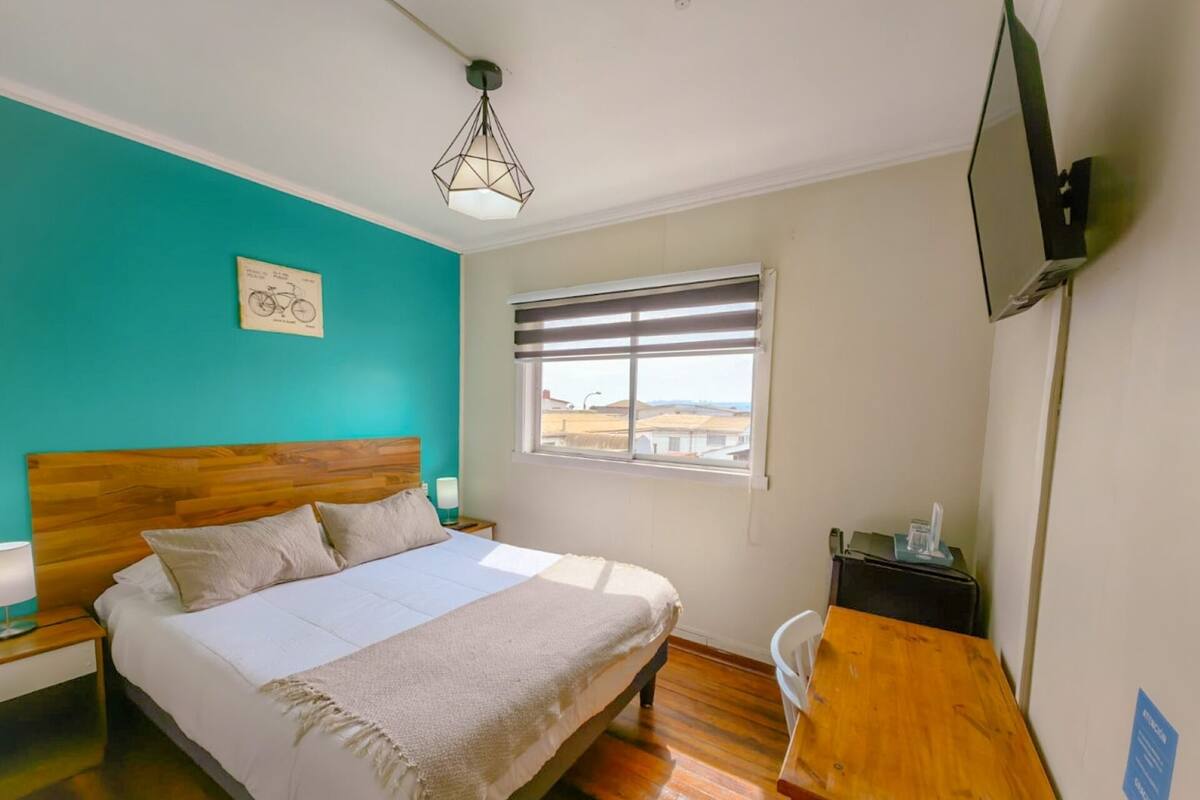 Un dormitorio con cama de madera, un escritorio, una silla, una ventana con persianas y un televisor instalado en la pared.