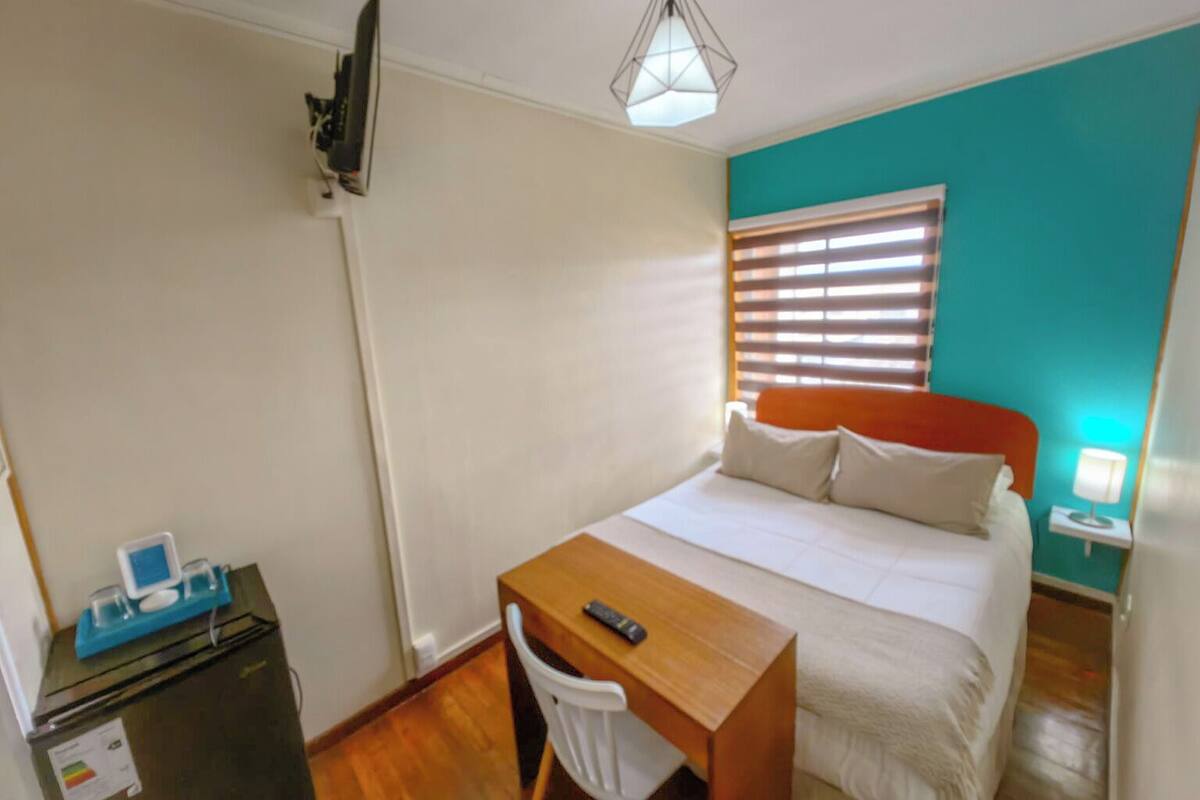 Un dormitorio con cama, escritorio, silla, una mesita con computadora y ventana con persianas.