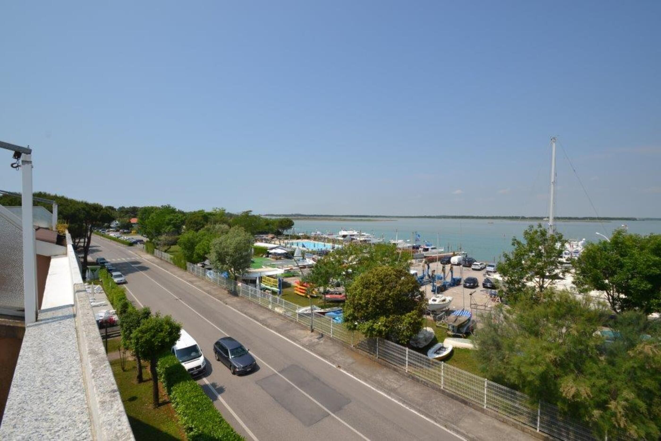 Una carretera costera con coches aparcados, un puerto deportivo con barcos y una vista despejada del mar.