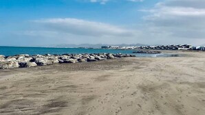 Ein Strand mit einem steinernen Wellenbrecher, ruhiger See und Gebäuden in der Ferne.