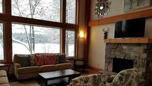 Ein gemütliches Wohnzimmer mit Kamin aus Stein, einem großen Fenster mit Blick auf eine verschneite Landschaft, einer Couch mit gemusterten Kissen und einem Fernseher, der über dem Kamin angebracht ist.
