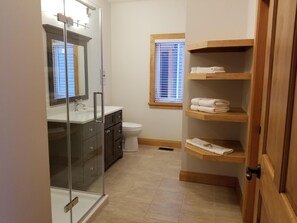Ein modernes Badezimmer mit Duschkabine aus Glas, einem weißen Waschbecken, dunklen Schränken, einer Toilette und Holzregalen mit gefalteten Handtüchern.