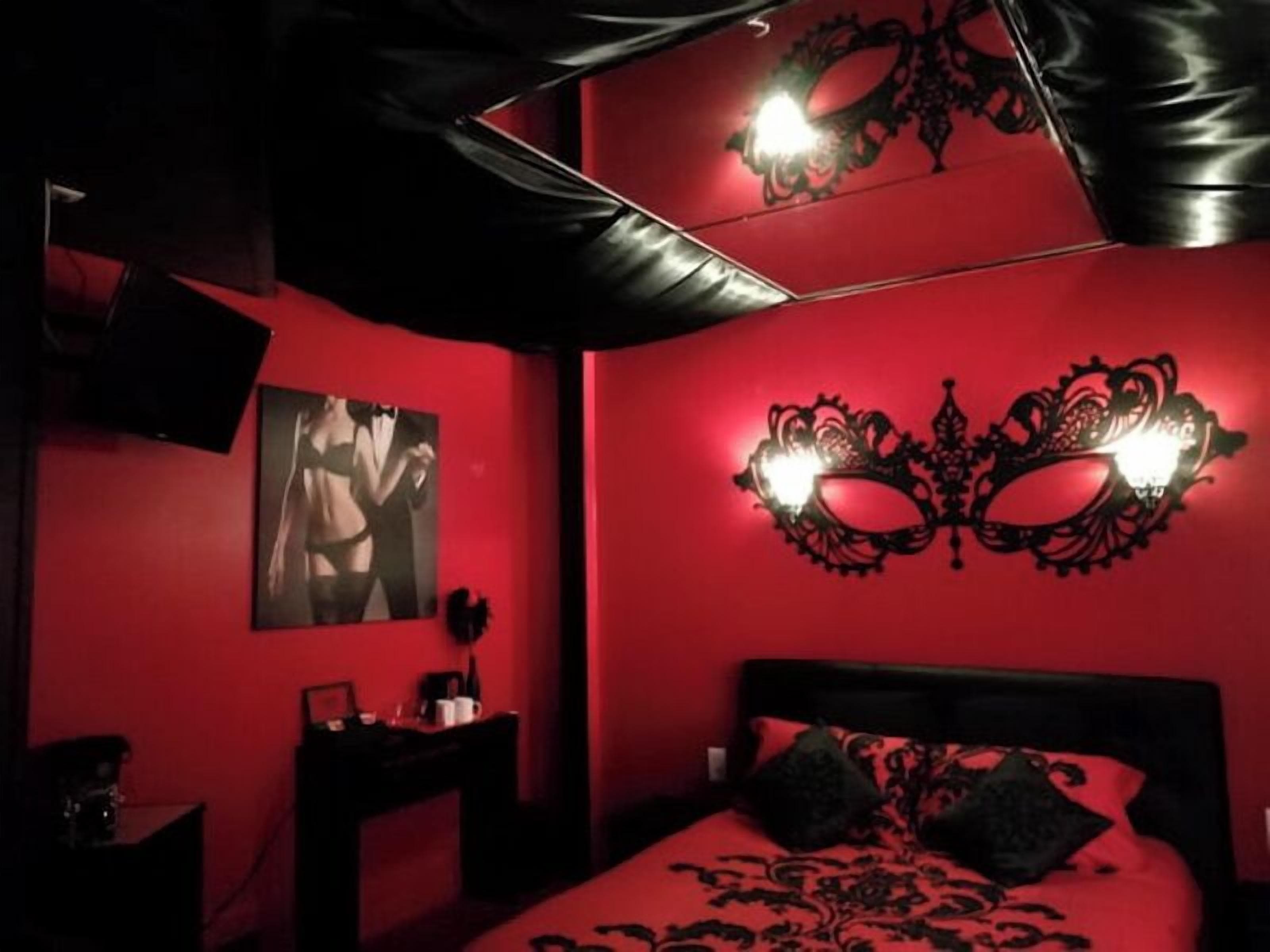 A room with a red wall, a bed with black bedding, a black headboard, a wall-mounted light fixture, and a framed picture on the wall.
