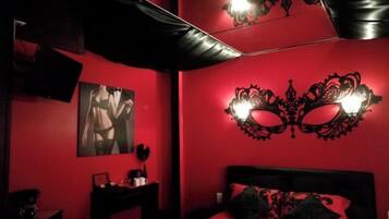 A room with a red wall, a bed with black bedding, a black headboard, a wall-mounted light fixture, and a framed picture on the wall.