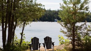Two Adirondack chairs facing a lake with a forested shoreline and a house in the background.