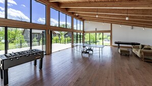 A spacious room with a foosball table, ping pong table, and a pool table. Large windows offer a view of the outdoors.