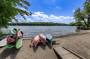 Un muelle en la orilla de un lago con kayaks y una tabla de remo, rodeado de árboles y un cielo despejado.