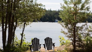 Two Adirondack chairs facing a lake with a forested shoreline and a house in the background.