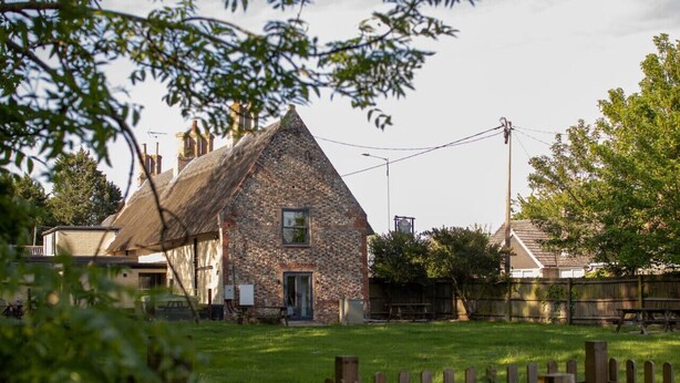A stone building with a thatched roof, surrounded by a wooden fence and a grassy yard. - Breckland Thatched Cottage: Hot Tub, 8 Cozy Rooms, Spacious Lounge (Hockwold cum Wilton)