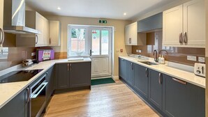 A modern kitchen with grey cabinets, a black stove, and a window with a view of the outside. - Breckland Thatched Cottage: Hot Tub, 8 Cozy Rooms, Spacious Lounge (Hockwold cum Wilton)