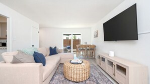 Un salon avec un canapé, un pouf, une télévision et, au fond, un coin repas.