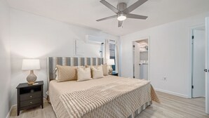 Une chambre à coucher comprenant un lit, une table de chevet, un ventilateur de plafond et une porte donnant sur une salle de bain.