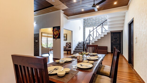 A dining area with a long wooden table set for six, flanked by cushioned chairs. A staircase with a metal railing is visible in the background. - Rahta Villa - An Elegant 3BR Villa With BBQ Terrace (Dehradun)