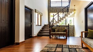 A spacious hallway with wooden flooring, a patterned rug, a staircase with a stone wall, and a seating area with a sofa and chairs. - Rahta Villa - An Elegant 3BR Villa With BBQ Terrace (Dehradun)