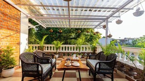 A covered patio with a checkered floor, two wicker chairs, a wooden table with plates and a pitcher, and hanging planters. - Rahta Villa - An Elegant 3BR Villa With BBQ Terrace (Dehradun)