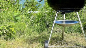 A black barbecue grill with a metal stand, positioned in front of a lush green area with a view of the ocean and a rocky coastline in the background. - 🌊 THE LOOKOUT -Ocean Views & Northern Lights (Kodiak)