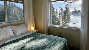 A bedroom with a bed, a bedside lamp, and two windows offering a view of a forest and a body of water. - 🌊 THE LOOKOUT -Ocean Views & Northern Lights (Kodiak)