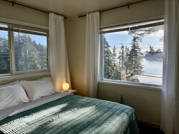 Un dormitorio con cama, una lámpara de noche y dos ventanas que ofrecen vista a un bosque y una masa de agua.