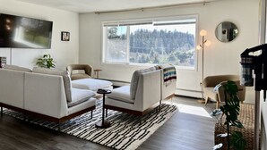 A modern living room with a sofa, armchairs, a coffee table, a TV, a large window with a forest view, and a floor lamp. - 🌊 THE LOOKOUT -Ocean Views & Northern Lights (Kodiak)