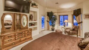 A spacious living room with a large brown carpet, wooden furniture, a flat-screen TV, and a window with curtains. - Havasu Jamaica S - Pool, Spa, Casita, Central (Lake Havasu City)
